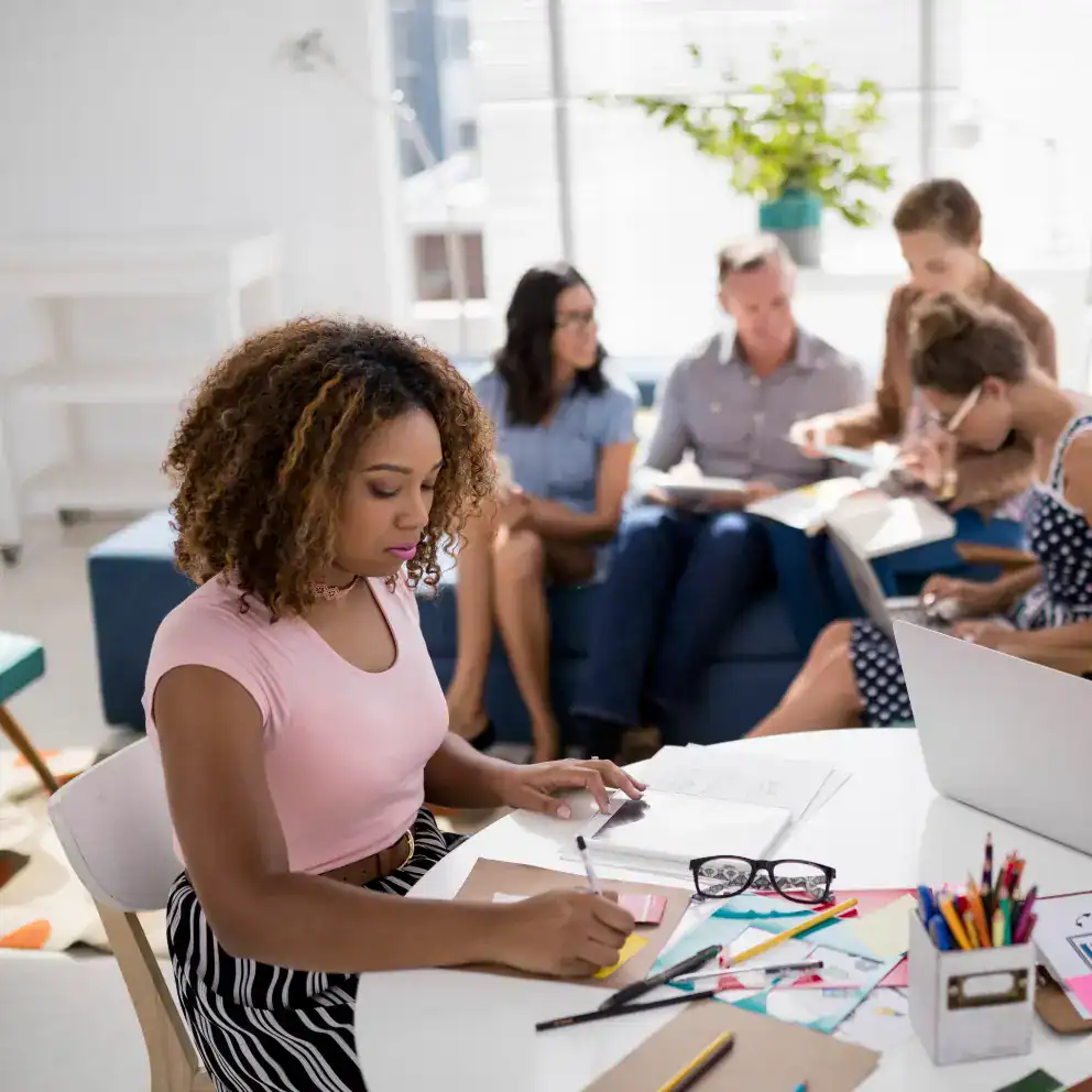 A woman works at a desk with papers and stationery, while a group of people have a discussion in the background in a bright, modern office.
