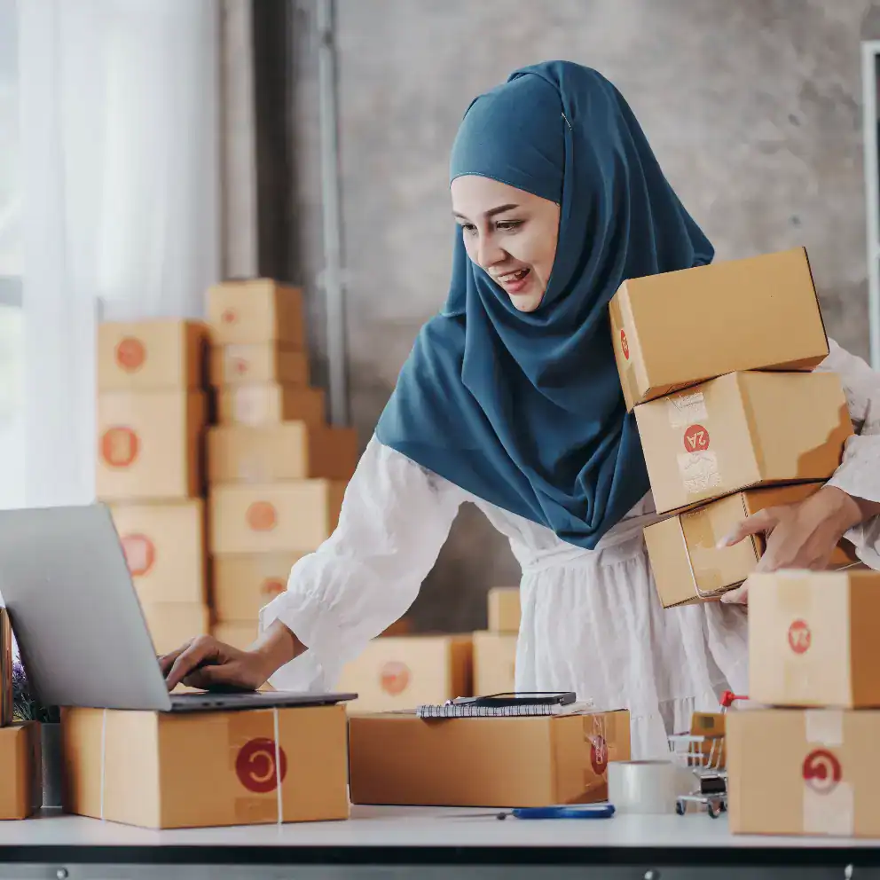 Woman wearing a blue hijab stands at a desk with several cardboard boxes, holding some in one arm and typing on a laptop with the other hand.