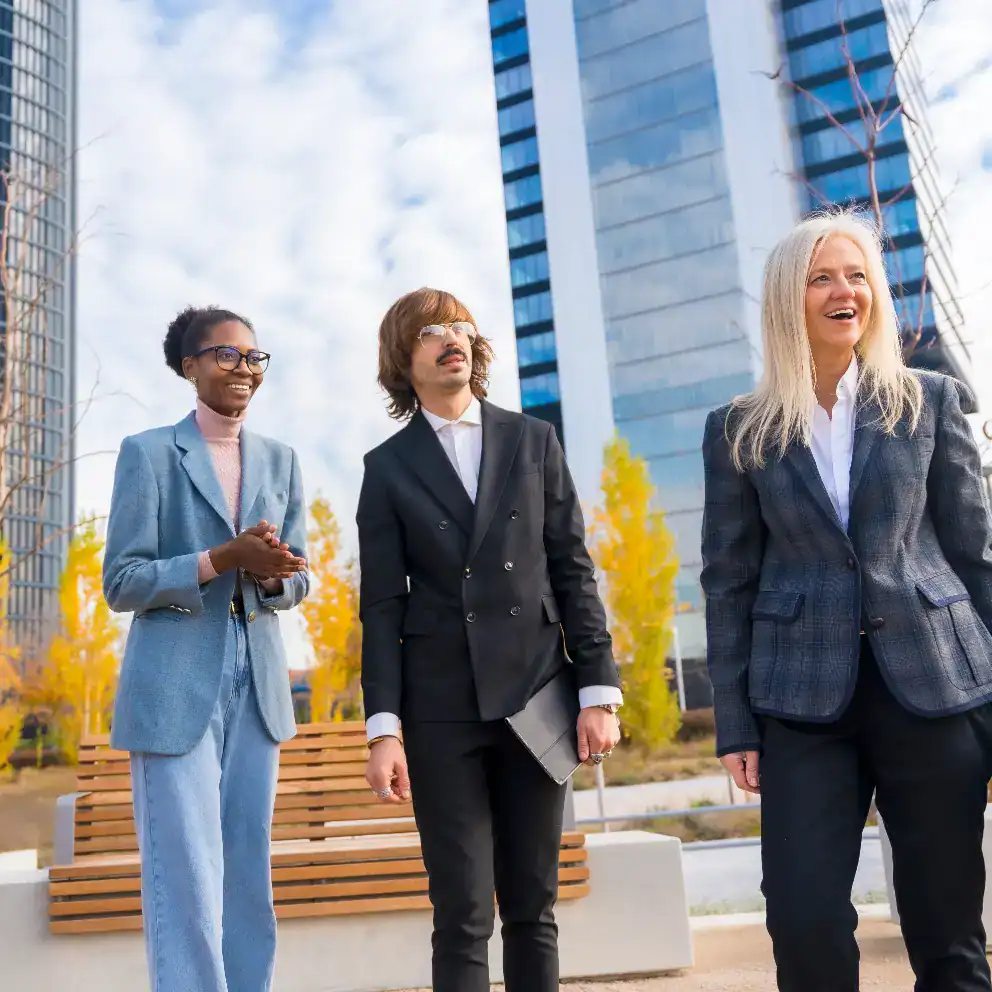 Three people in business attire stand outdoors in front of modern office buildings, smiling and talking on a clear day.