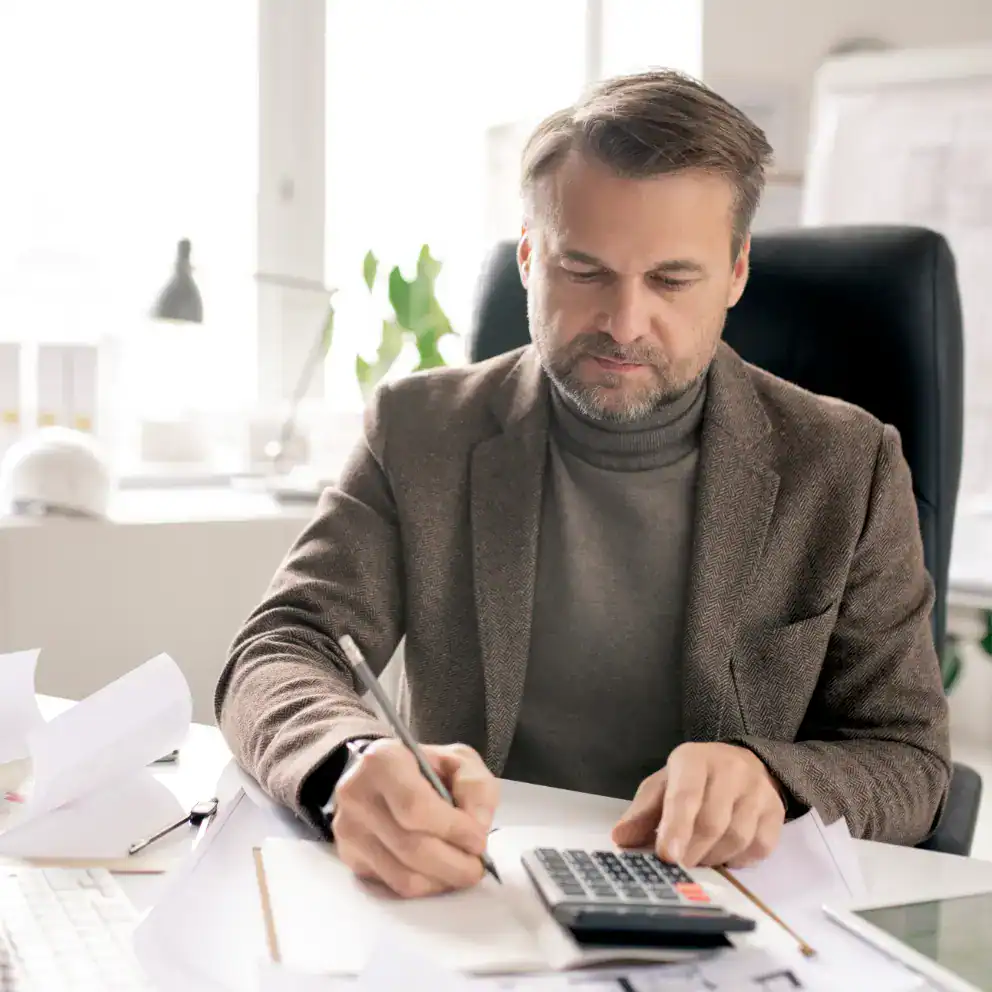 Man in a suit sits at a desk using a calculator and writing on paperwork, surrounded by documents in a bright office setting.