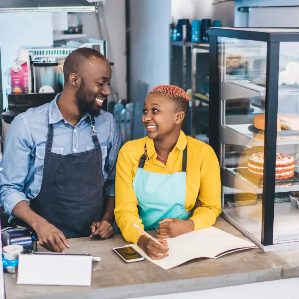 Two people wearing aprons stand behind a bakery counter, smiling at each other, with a display case of cakes and an open notebook nearby.