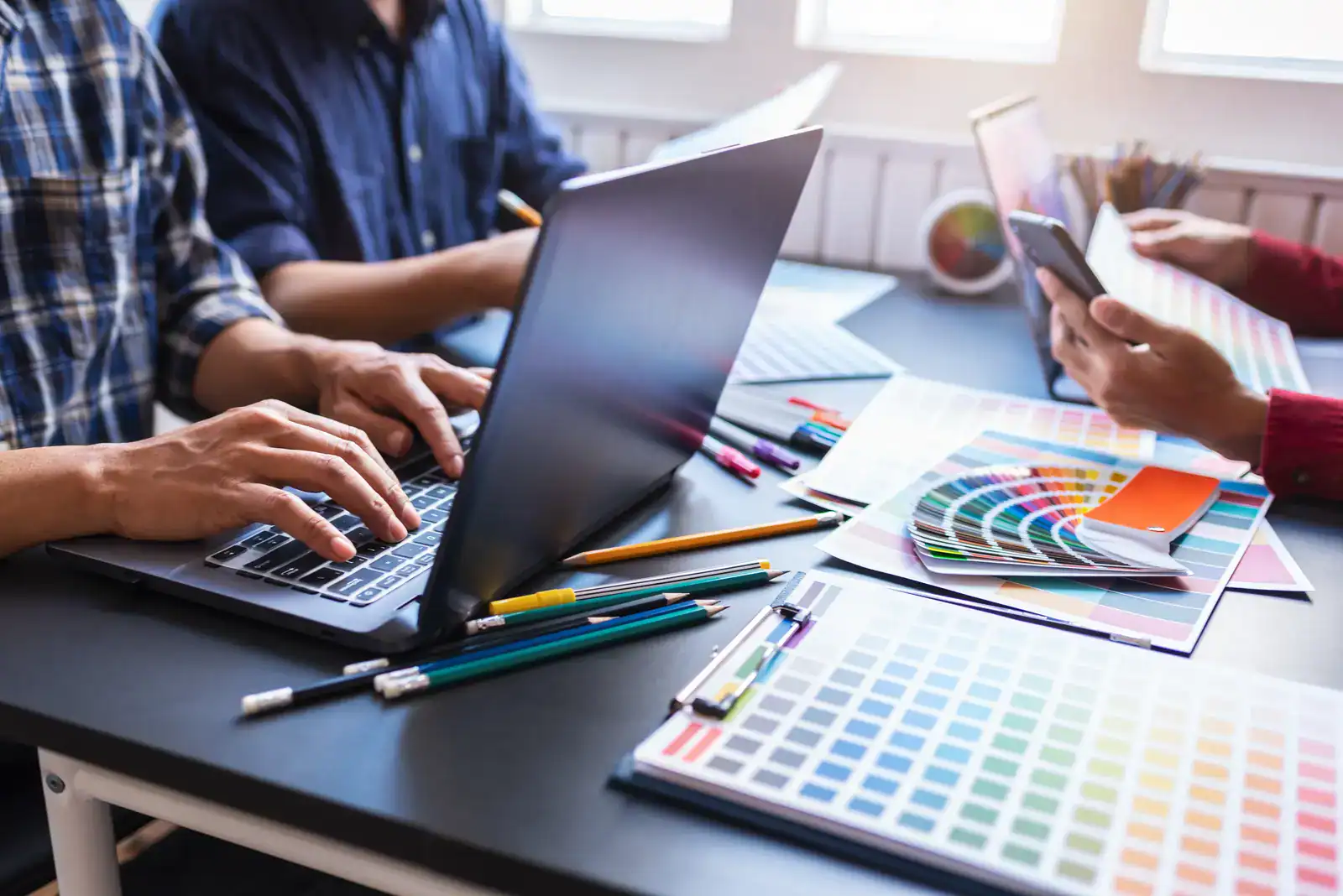 Three people work at a desk with a laptop, smartphone, pencils, and colour swatches, reviewing design samples and palettes.