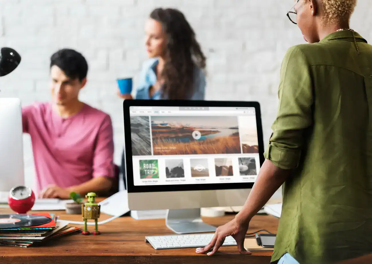 Three people work in a modern office; one stands at a desk using a computer with a travel website on the screen, while two others talk in the background.
