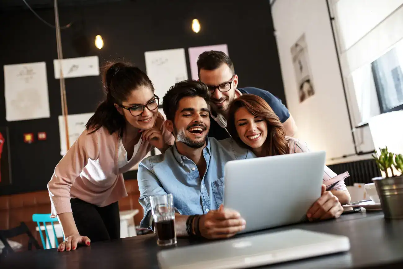 Four people sit and stand around a table, smiling and looking at a laptop screen together in a casual office setting.