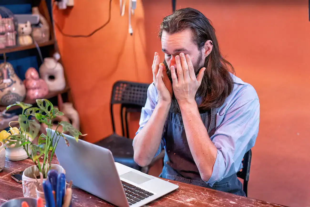 A person wearing an apron sits at a table with a laptop, covering their face with their hands, appearing stressed or tired in a workspace.
