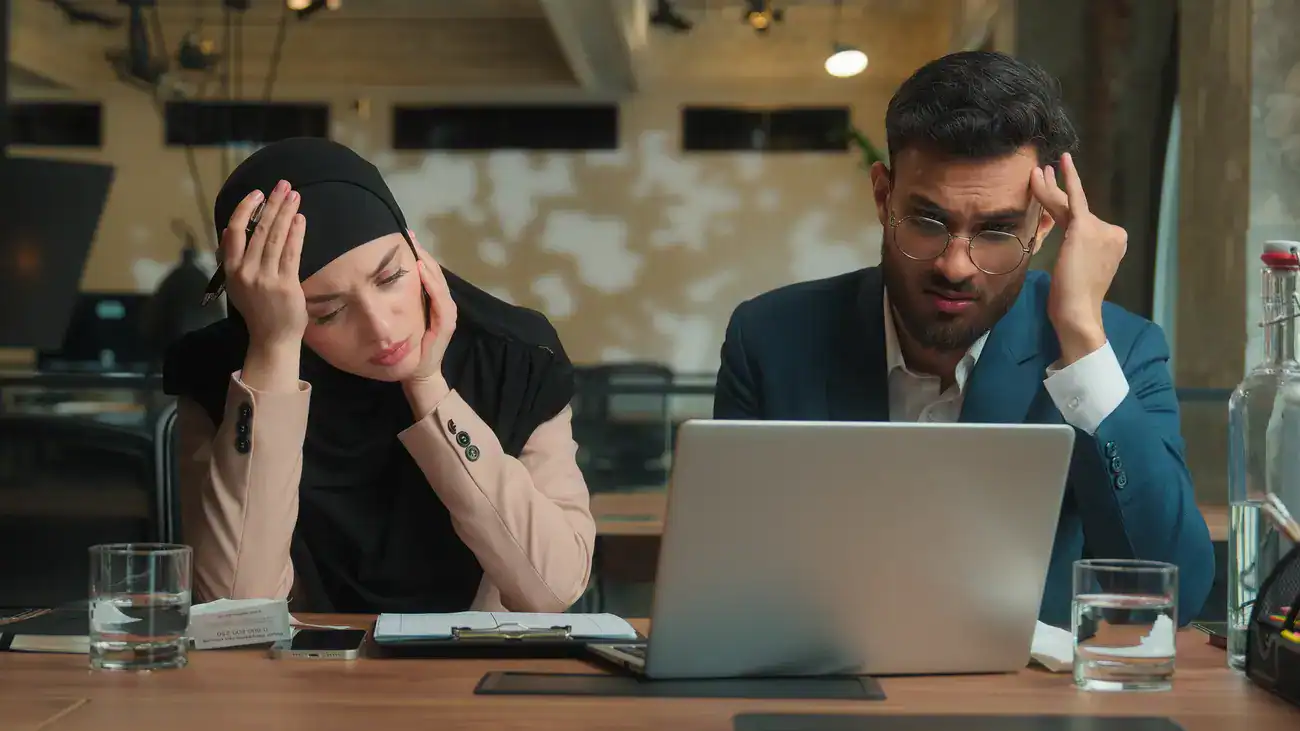 Two people sit at a desk looking stressed while facing a laptop, with notepads, water glasses, and papers in front of them in an office setting.