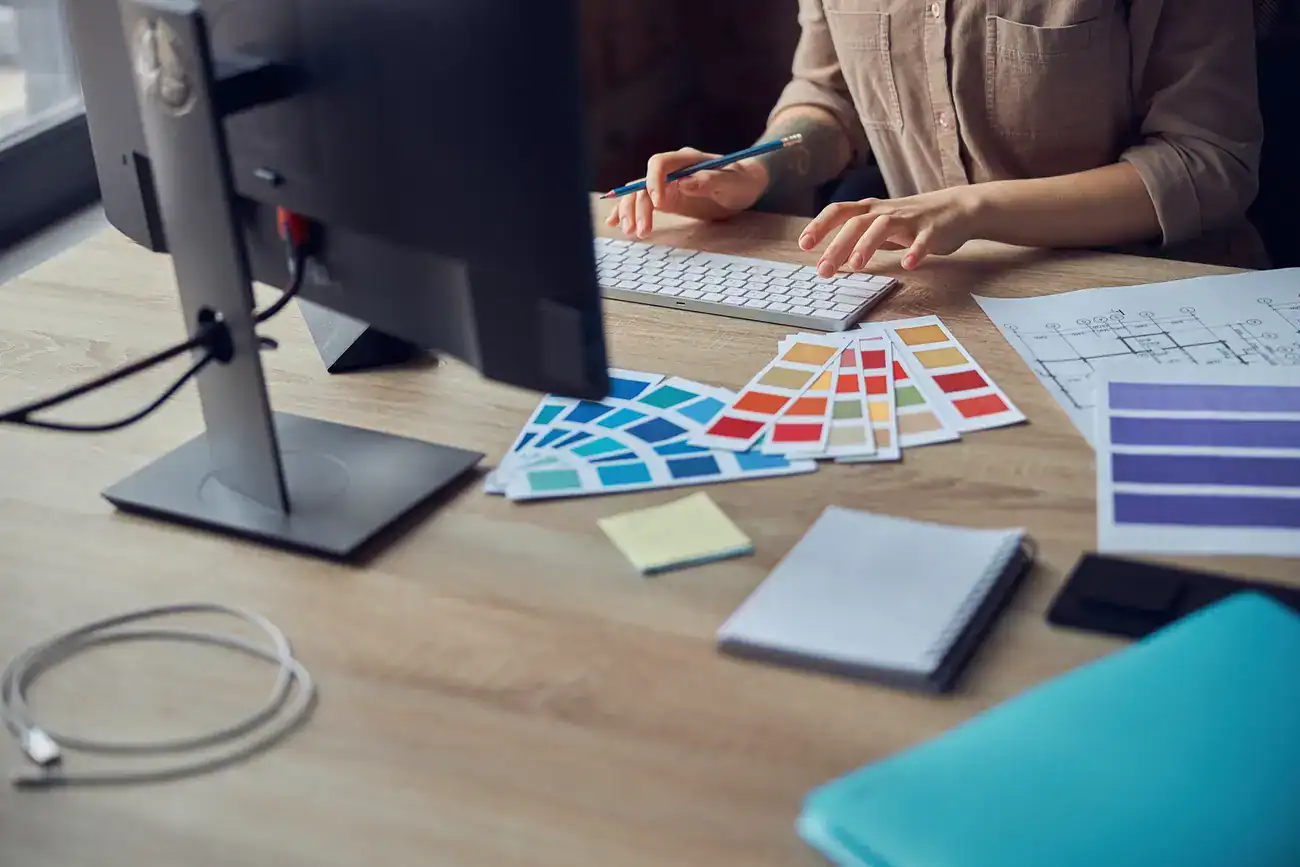 Person working at a desk with a computer, colour swatches, papers, a notebook, sticky notes, and a blue folder.