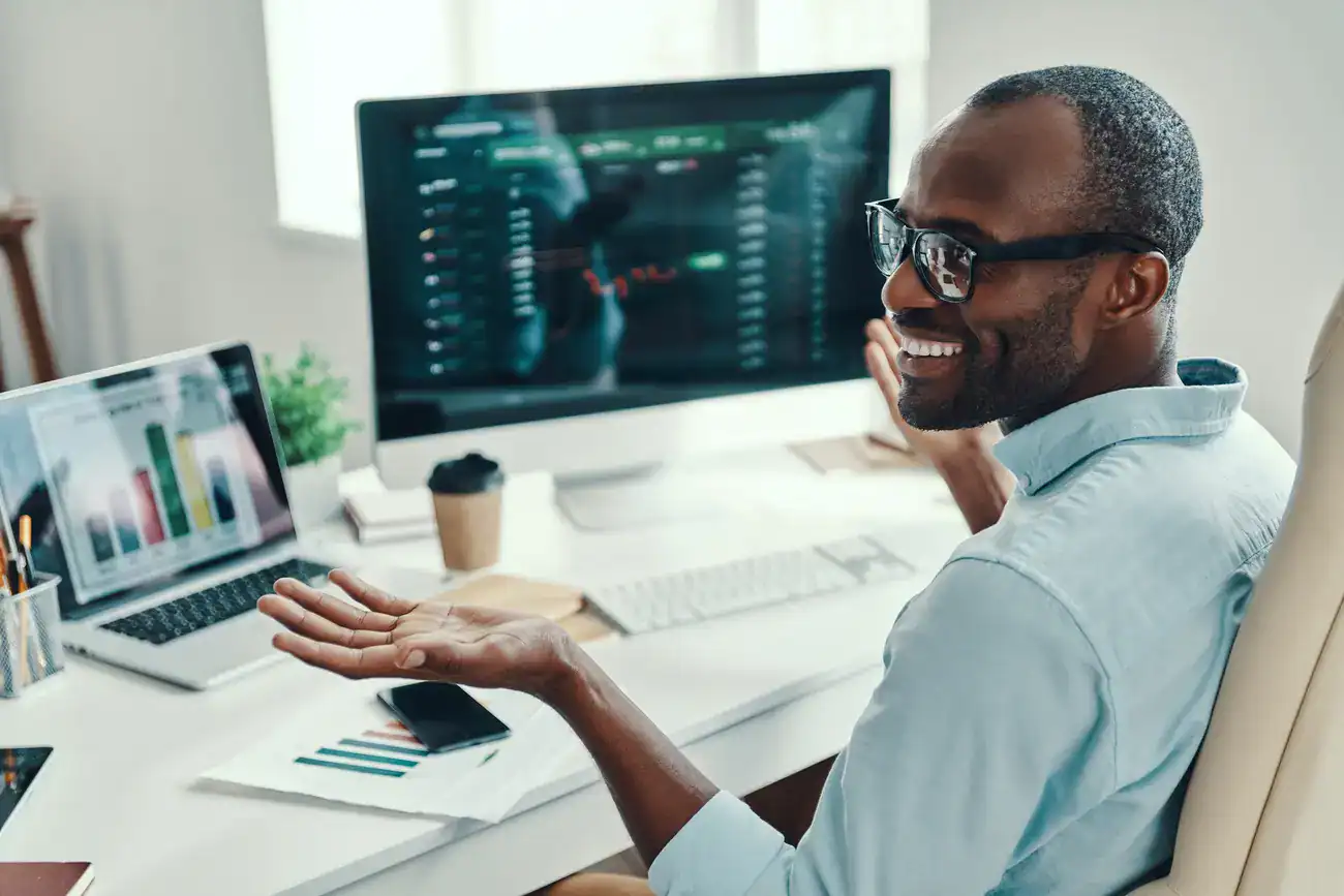 A man wearing glasses sits at a desk with graphs and charts on computer screens, gesturing with one hand and smiling.