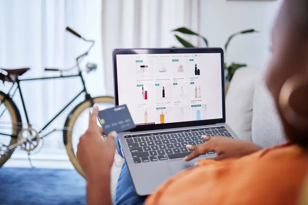 Person holding a credit card while shopping online on a laptop, with product listings displayed on the screen and a bicycle in the background.