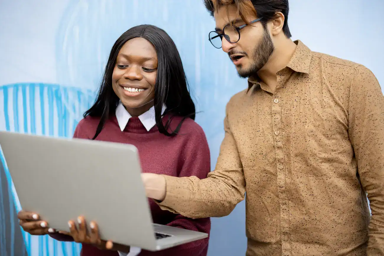 Two people stand together looking at a laptop; the woman holds the laptop and smiles while the man points at the screen and speaks.