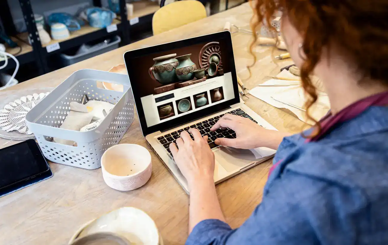 A person using a laptop to browse a pottery website at a table with ceramics, pottery tools, and a basket of supplies.