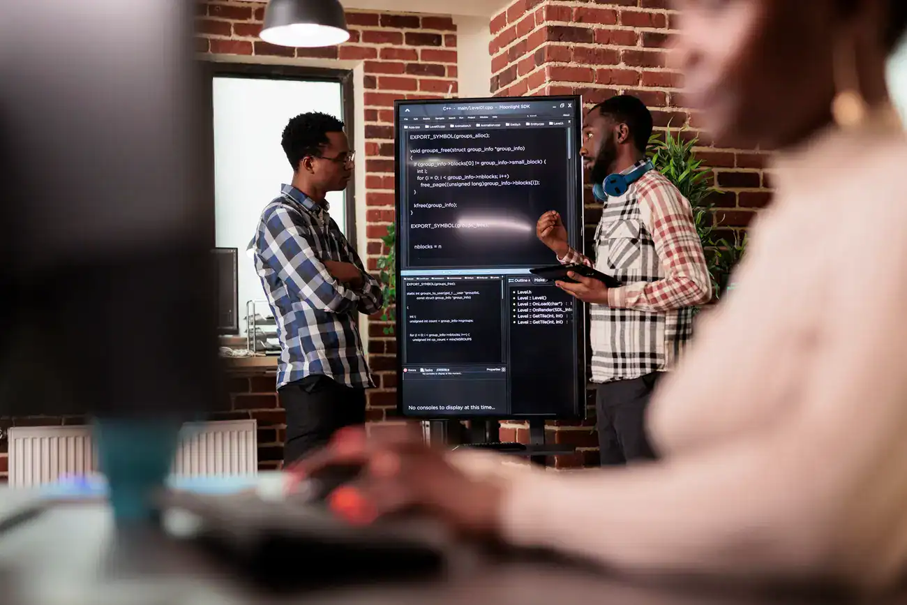 Two people discuss code displayed on a large screen in an office with brick walls, while another person works at a computer in the foreground.