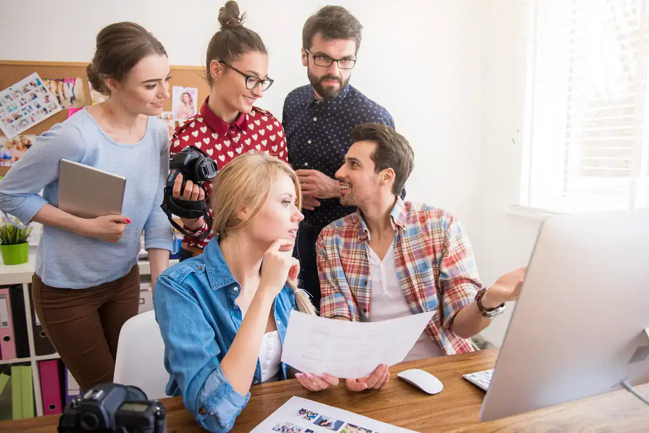 Five people collaborate around a desk with a computer, camera, and papers, appearing to discuss a creative project in a bright office space.