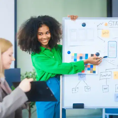 A woman in a green shirt presents information on a whiteboard covered with charts and diagrams while another person holds a tablet in the foreground.
