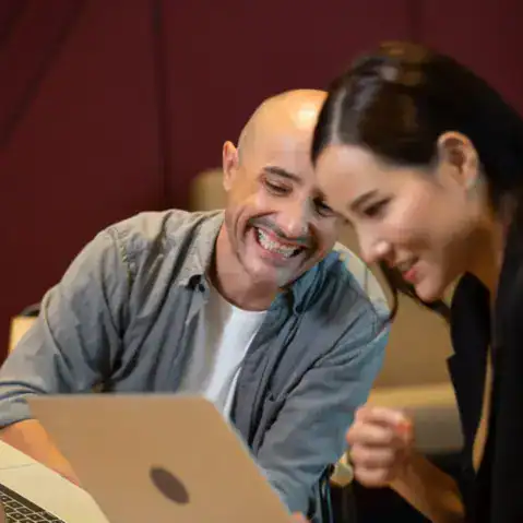A man and a woman sit together at a table, smiling and looking at a laptop screen.