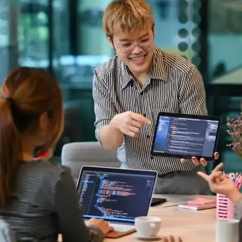 A person stands and shows code on a tablet to two colleagues, who are seated with laptops displaying code in an office meeting room.
