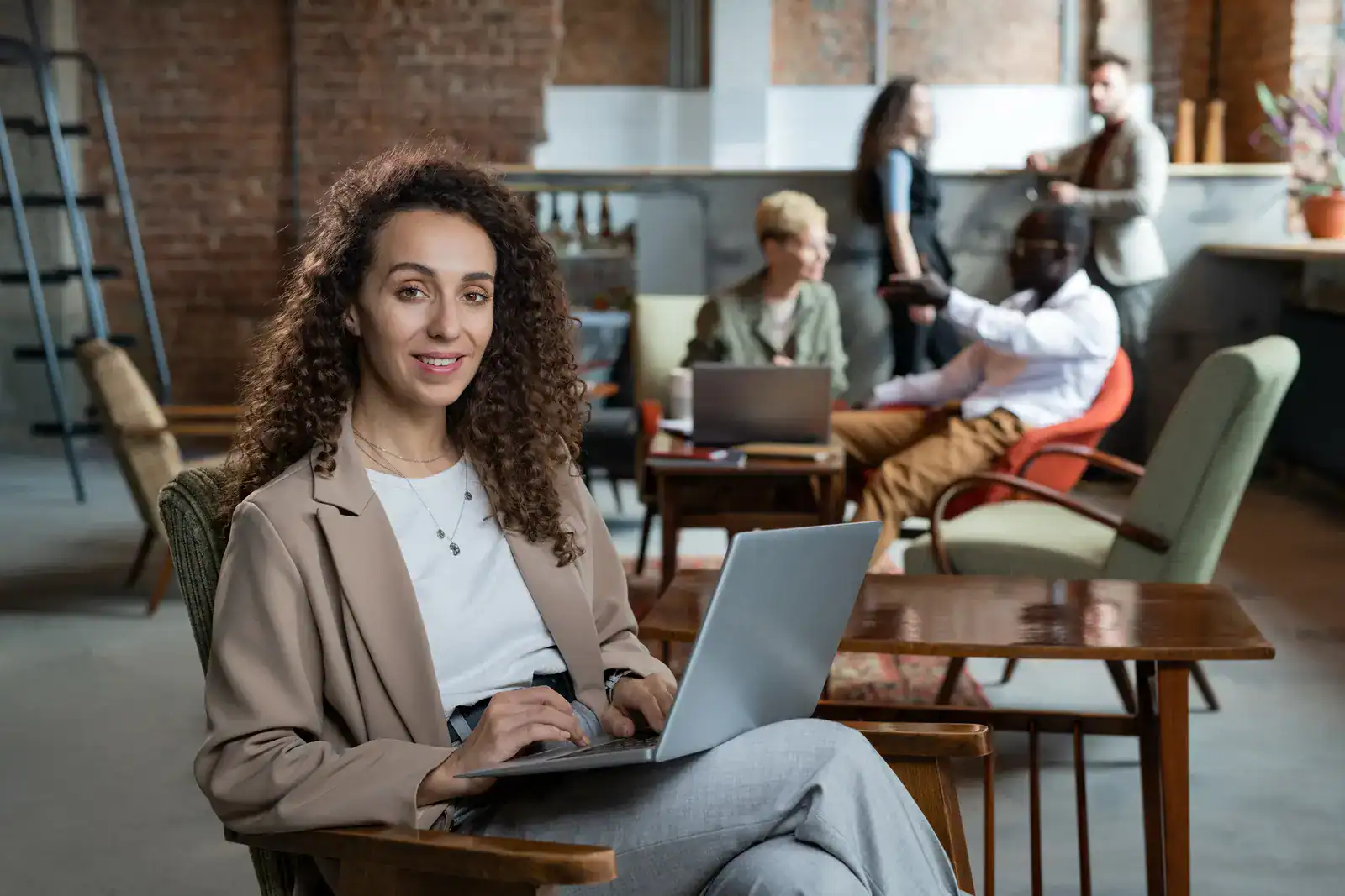 Woman with curly hair working on a laptop in a modern office lounge; three colleagues converse and work in the background.