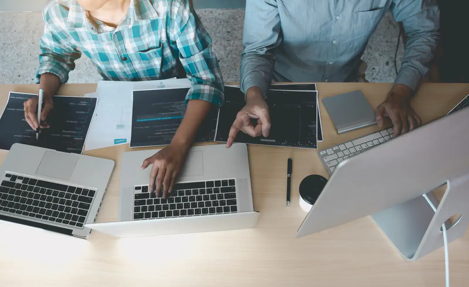 Two people work at a desk with laptops, documents, and a desktop monitor, collaborating and pointing at the screen.