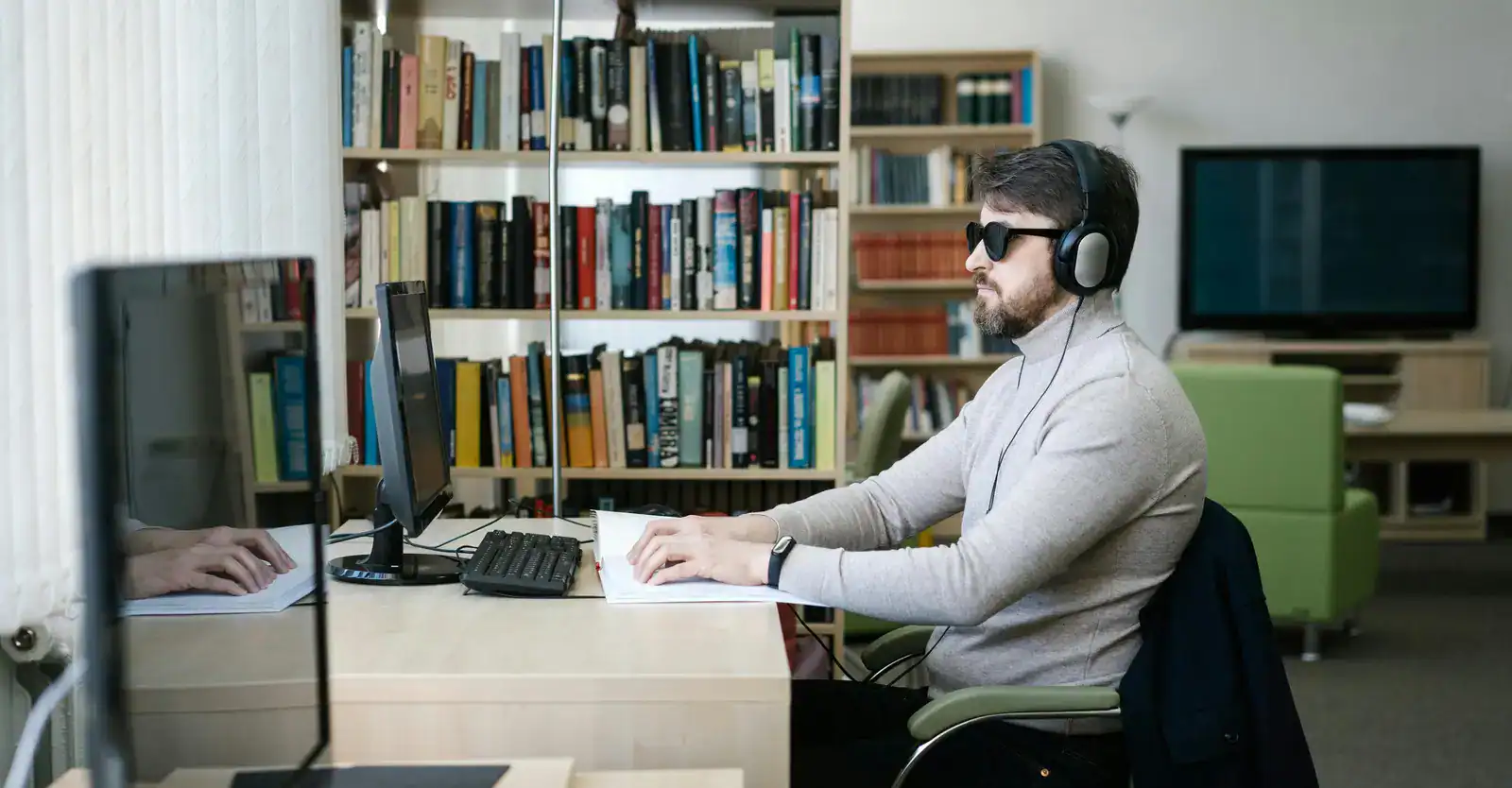 A man wearing sunglasses and headphones uses a computer and reads braille at a desk in a library or office setting.