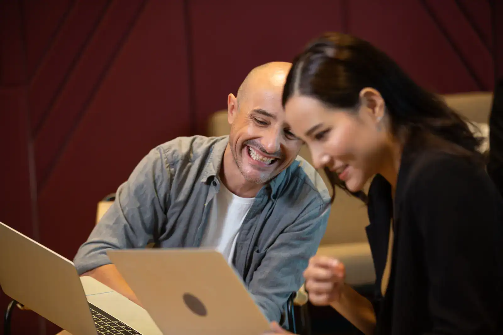 A man and a woman sit side by side, smiling and looking at a laptop screen together in an indoor setting.