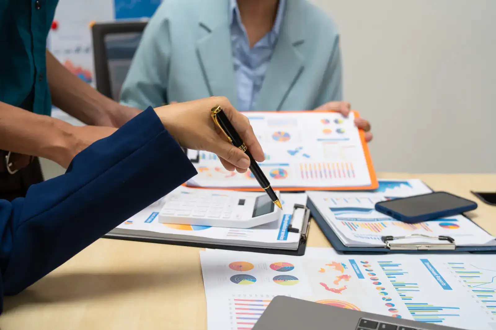 Three people in business attire review printed financial charts and graphs at a table with a calculator, clipboard, smartphone, and laptop.
