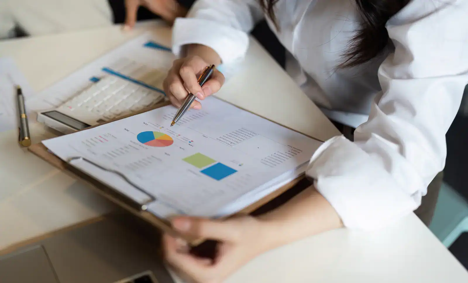A person reviews financial charts and graphs on a clipboard, holding a pen, with documents and a laptop on the desk.