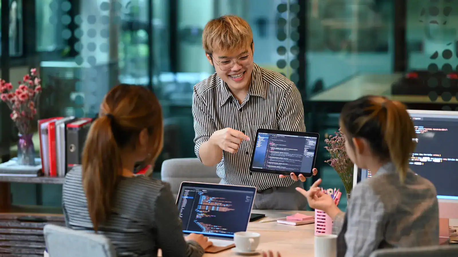 A person shows code on a tablet to two colleagues during a meeting; laptops with code are open on the table in a modern office setting.