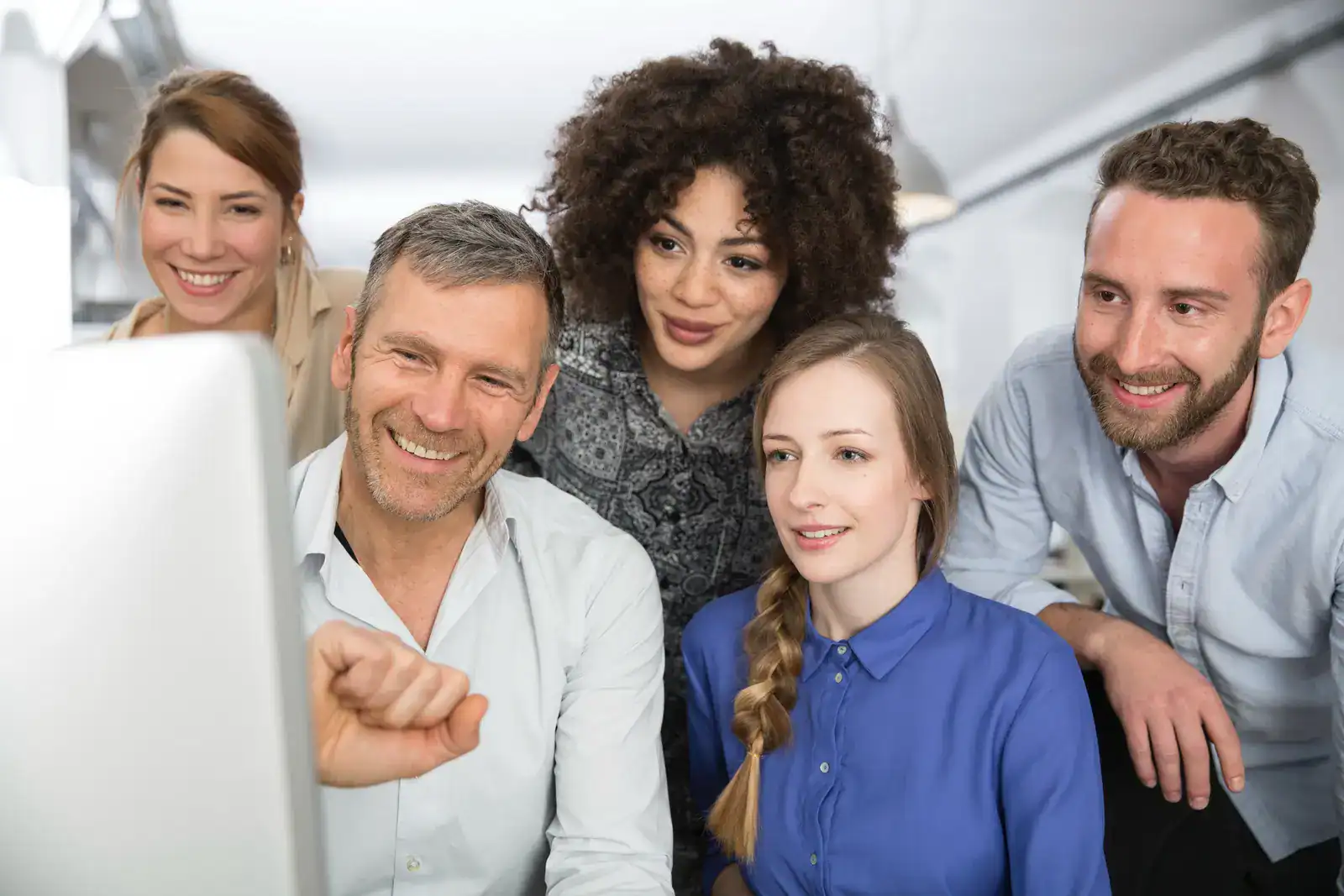 Five people, three women and two men, gather around a computer monitor, smiling and looking at the screen in a bright office setting.