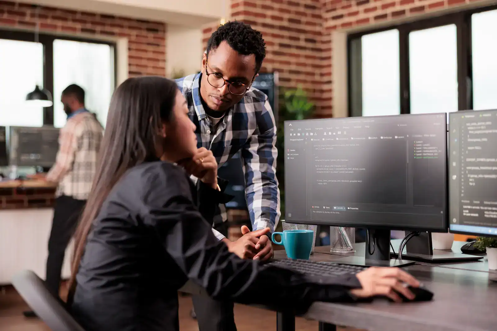 A man and woman discuss code displayed on a computer monitor in a modern office workspace with exposed brick walls.