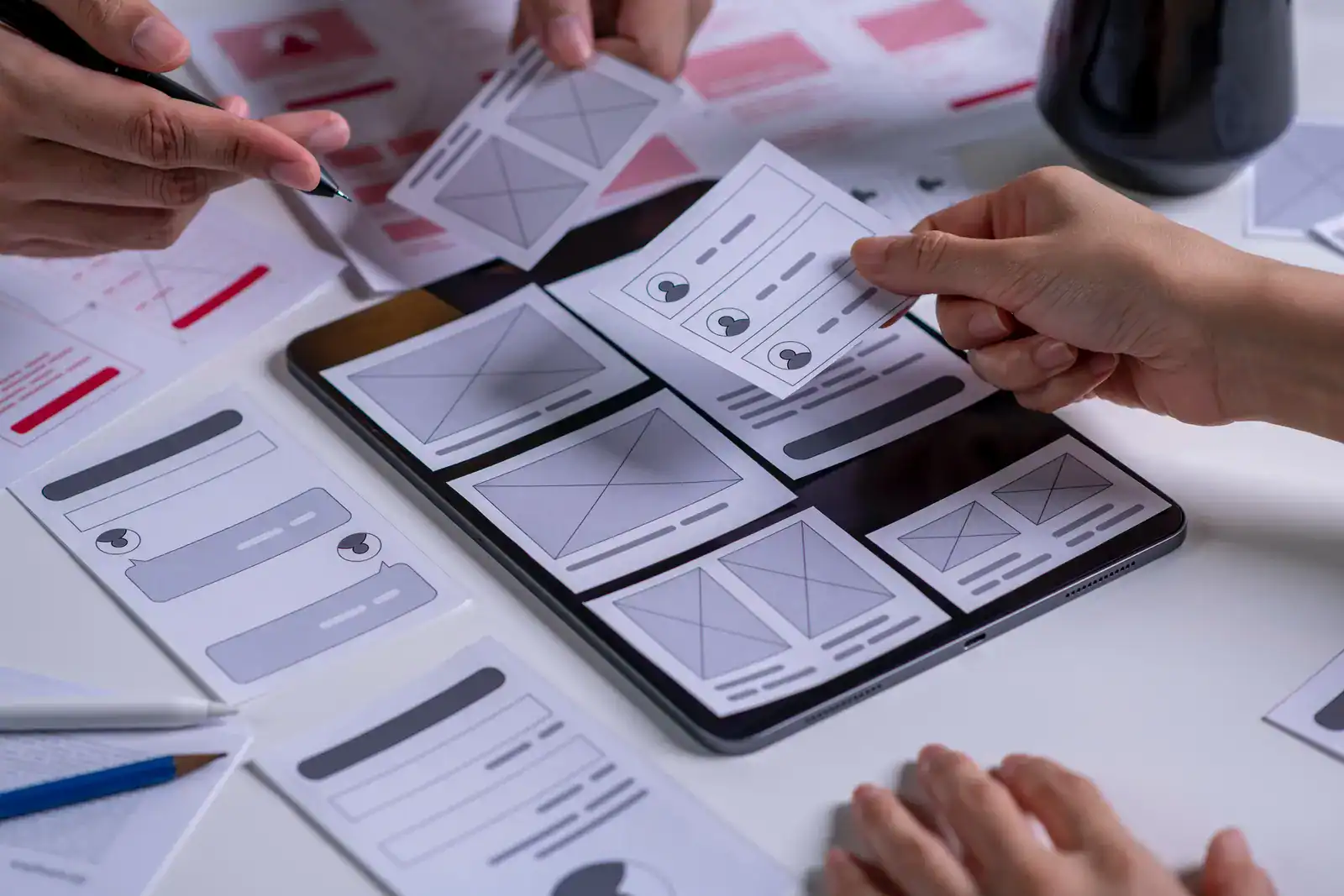 Hands arrange paper prototypes of website or app interface screens on a tablet, with additional design mockups and documents on a desk.