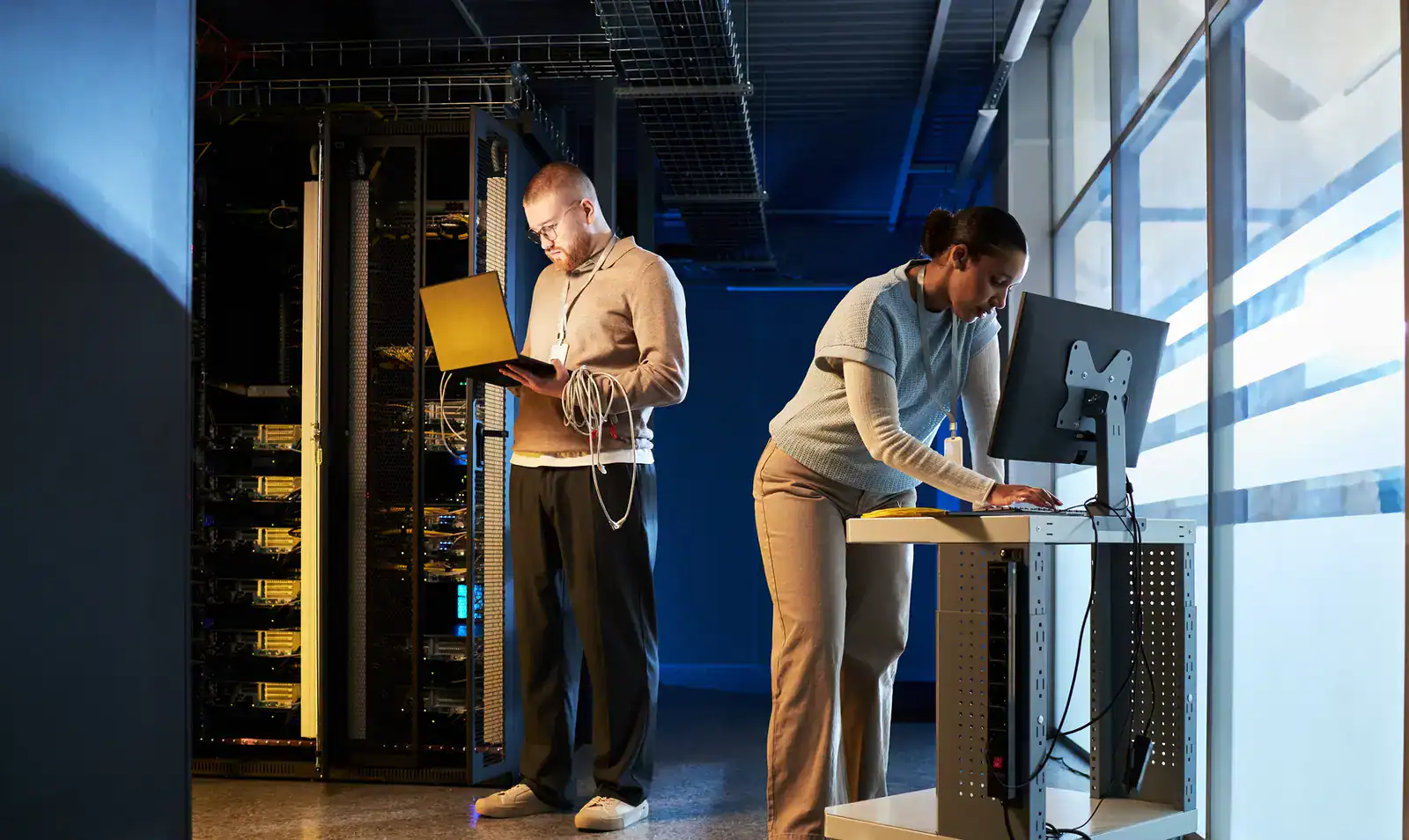 Two IT professionals work in a server room; one uses a laptop near server racks, while the other types on a desktop computer at a workstation.