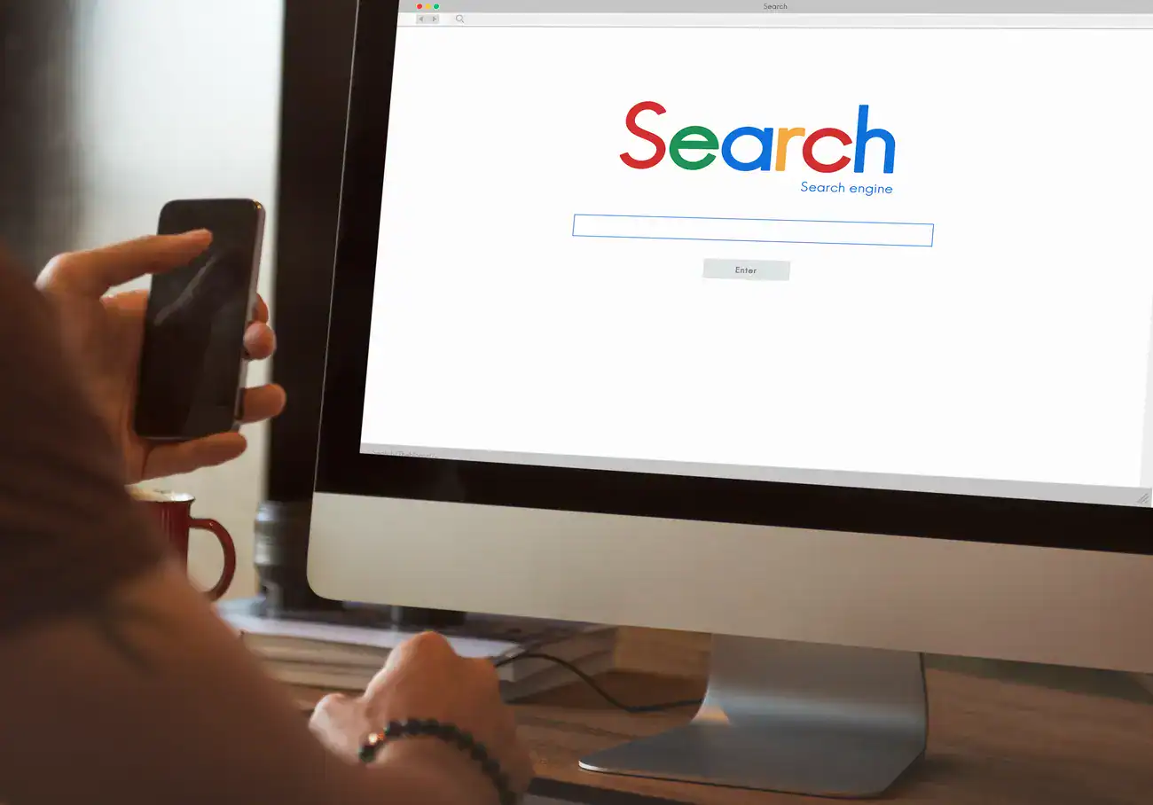 Person holding a smartphone and sitting at a desk in front of a computer displaying a search engine homepage with a search bar and "Search" written in colourful letters.