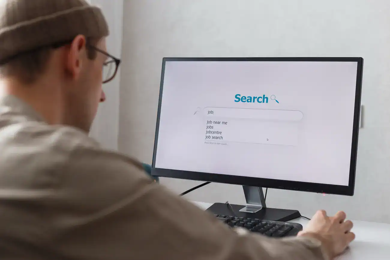 Person wearing glasses and a beanie using a desktop computer to search online, with search suggestions visible on the screen.