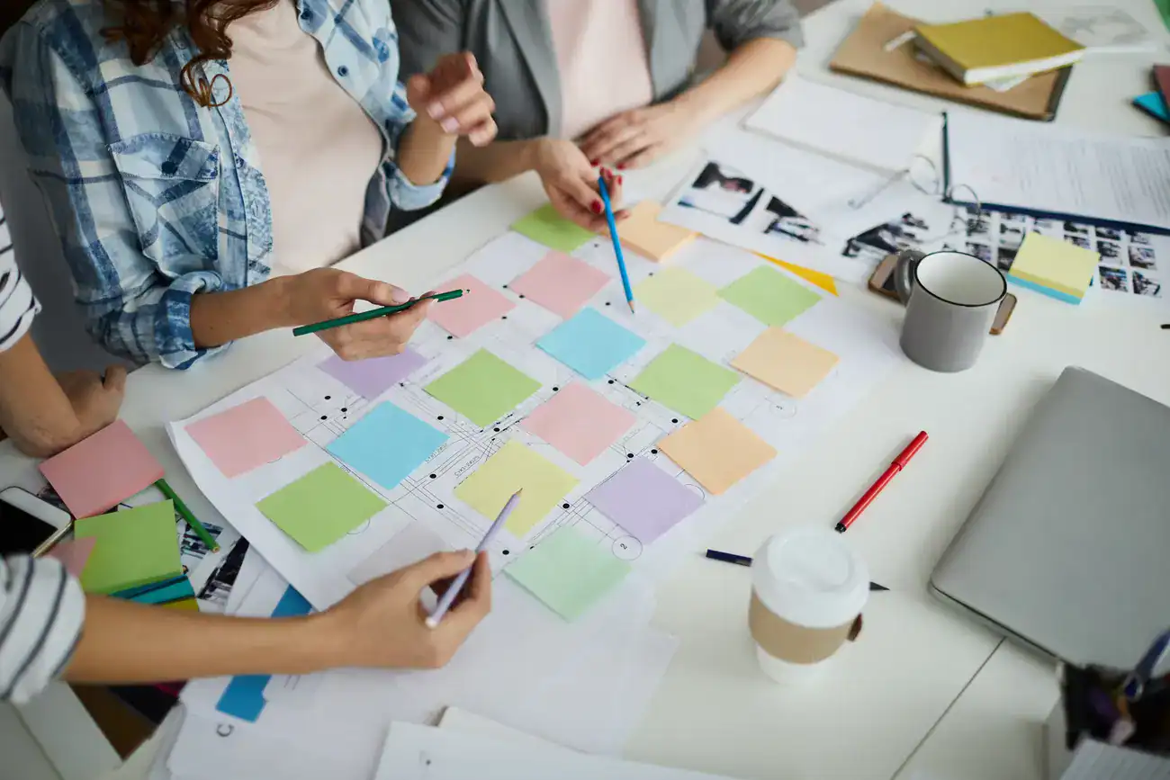 Three people collaborate at a table covered with colourful sticky notes, documents, pens, and drinks, appearing to plan or organize a schedule or project.