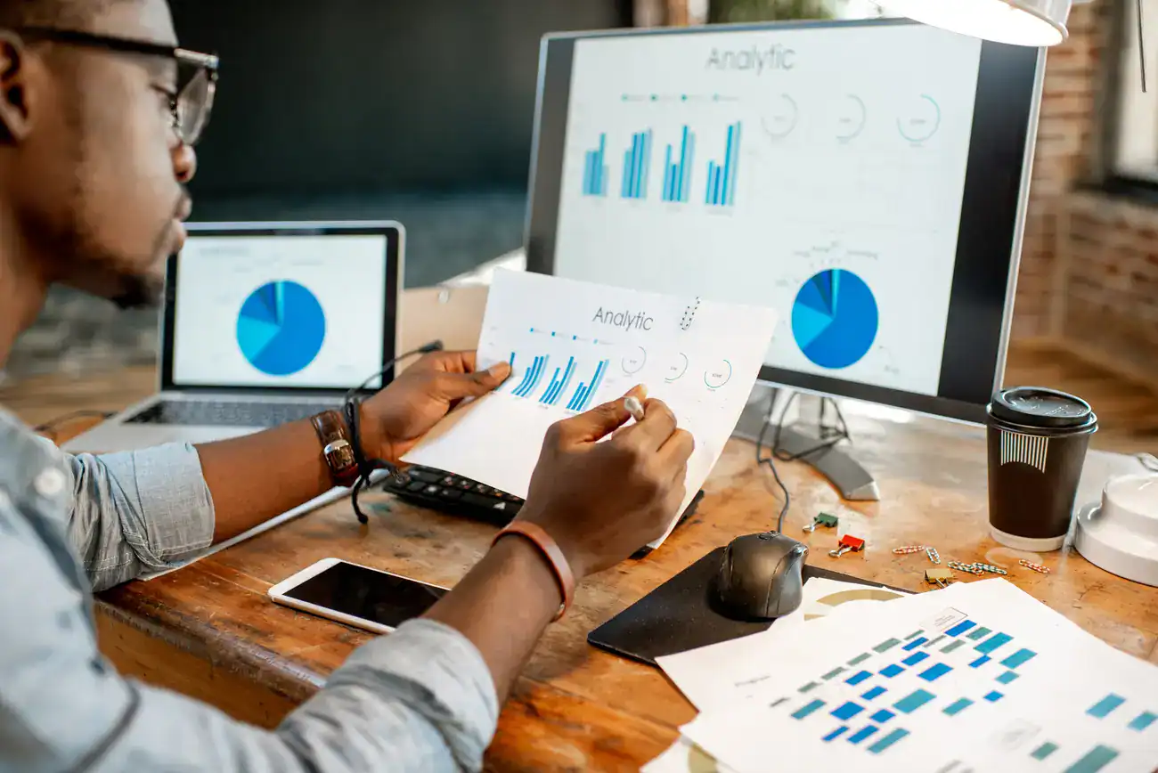 A marketer reviews competitor analytics on paper printouts, his laptop, and a secondary screen